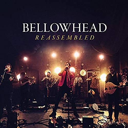 Bellowhead band members performing on a dimly lit stage with bright spotlights behind them.