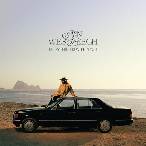 Man in hat sits on car with ocean and mountains backdrop.