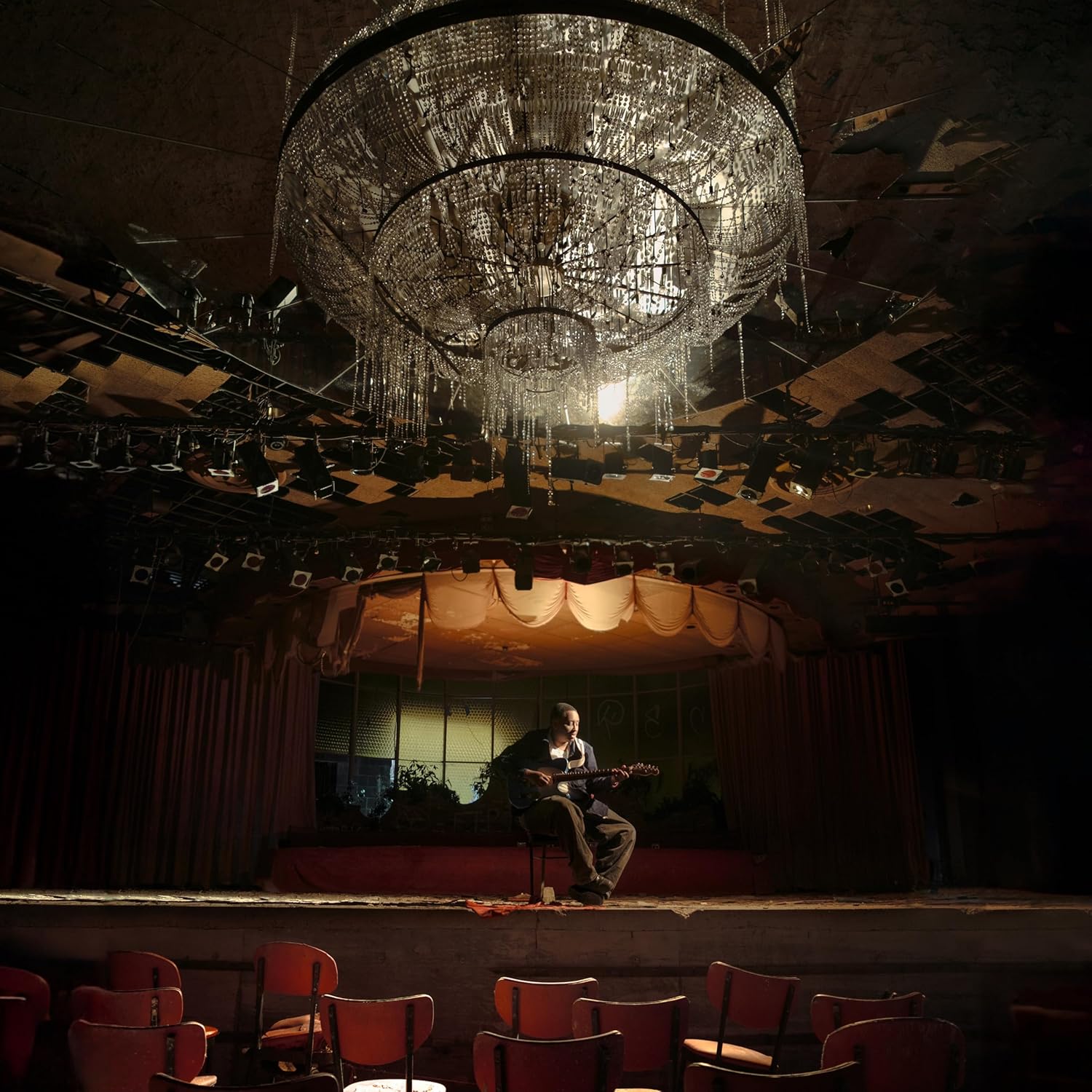 Musician playing guitar on a dimly lit, empty stage under a chandelier.