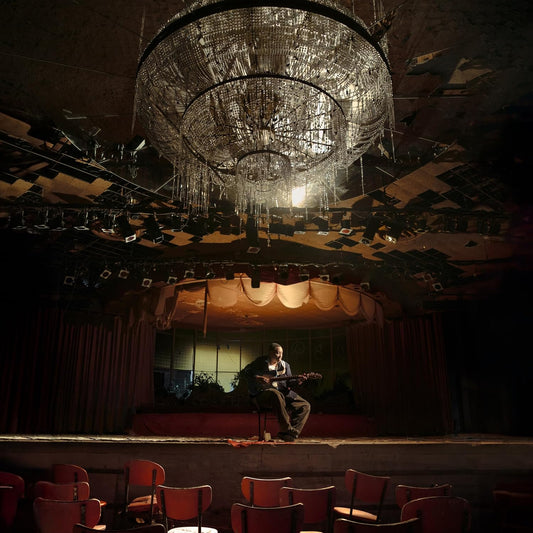 Musician playing guitar on a dimly lit, empty stage under a chandelier.