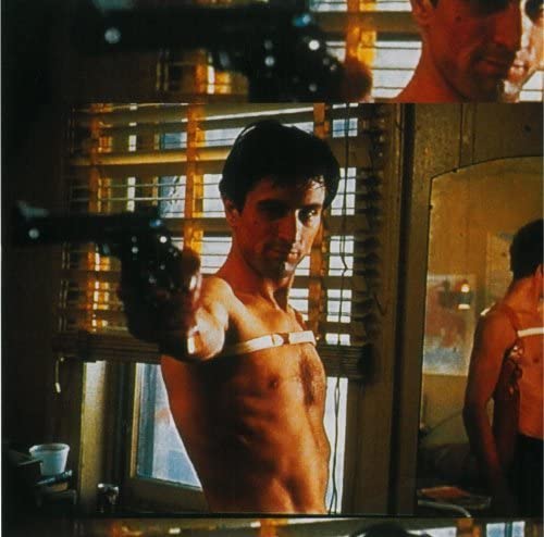 Robert De Niro shirtless, holding a handgun, reflected in a mirror with window blinds behind.
