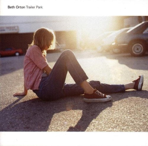 A woman sits on asphalt in a parking lot at sunset, wearing jeans and sneakers.
