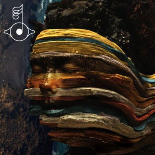 A layered, abstract face sculpture against a dark, rocky background, with an eye symbol.
