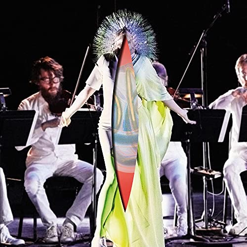 Björk stands center stage in a yellow gown before an orchestra in white clothing.