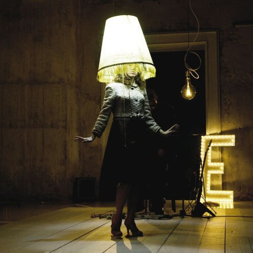 A woman wearing a lampshade on her head stands in a dimly lit room.