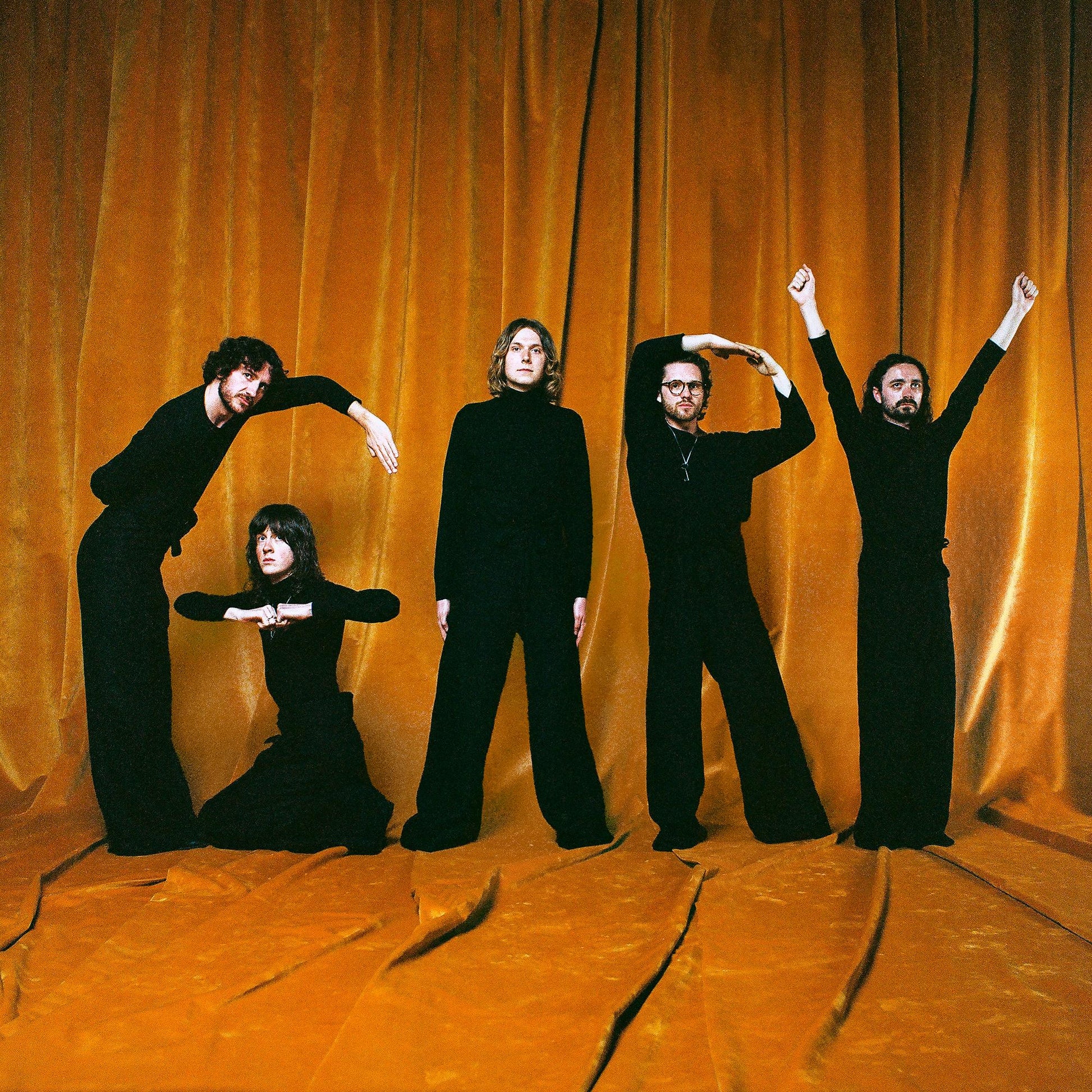 Four people in black clothing pose with raised arms against orange curtains.