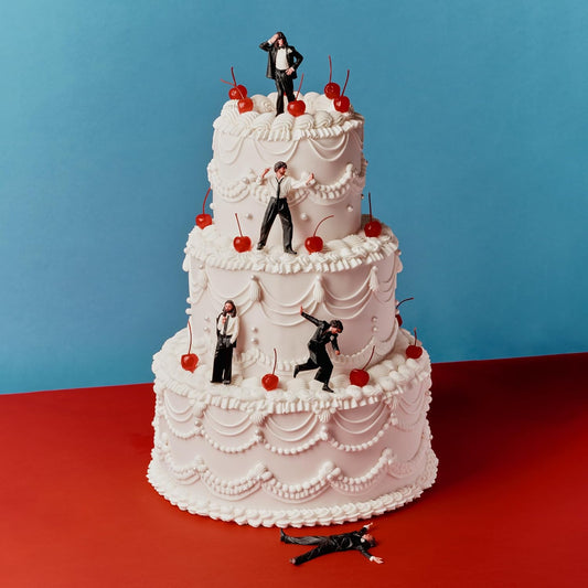 A three-tiered white wedding cake decorated with cherries and miniature figures of men.