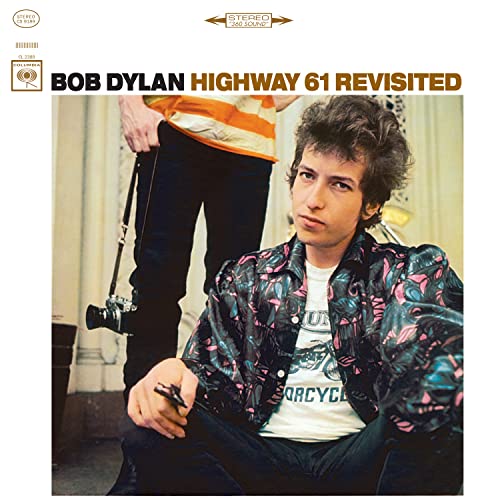 Bob Dylan sits in a patterned shirt with a person holding a camera behind him.