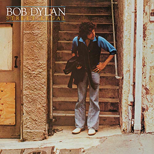 Bob Dylan standing on stone stairs holding a jacket, wearing jeans and blue shirt.