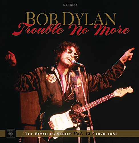 Bob Dylan performing on stage with a guitar, wearing sunglasses, arms raised, black background.