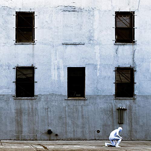 The image shows a vinyl record cover featuring a person crouching in front of a grey building.
