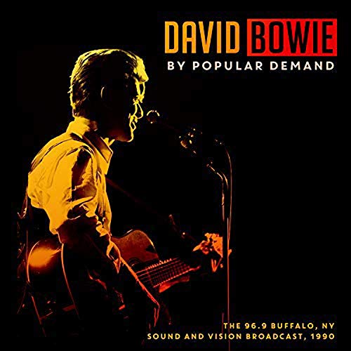 David Bowie performing on stage with an acoustic guitar, illuminated in orange against black.
