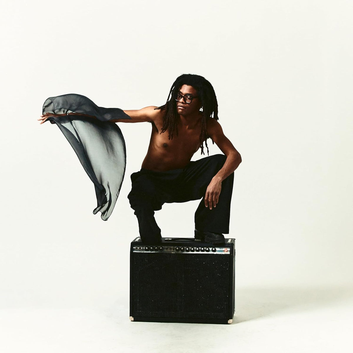 Man with dreadlocks squats on an amplifier, holding flowing fabric.