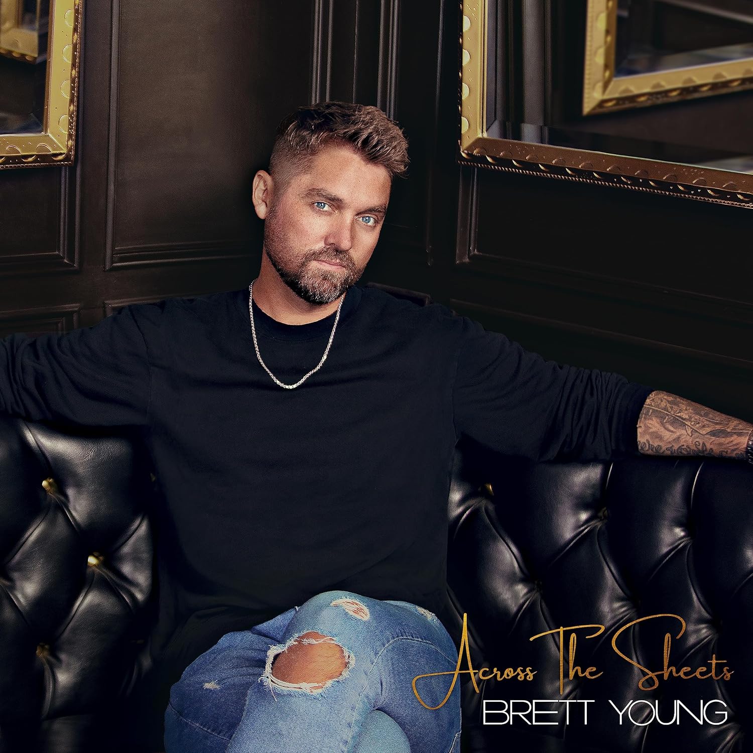 Brett Young sits on a black leather couch wearing a black shirt and ripped jeans.
