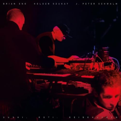 Three musicians perform with electronic equipment in a dimly lit room with red lighting.