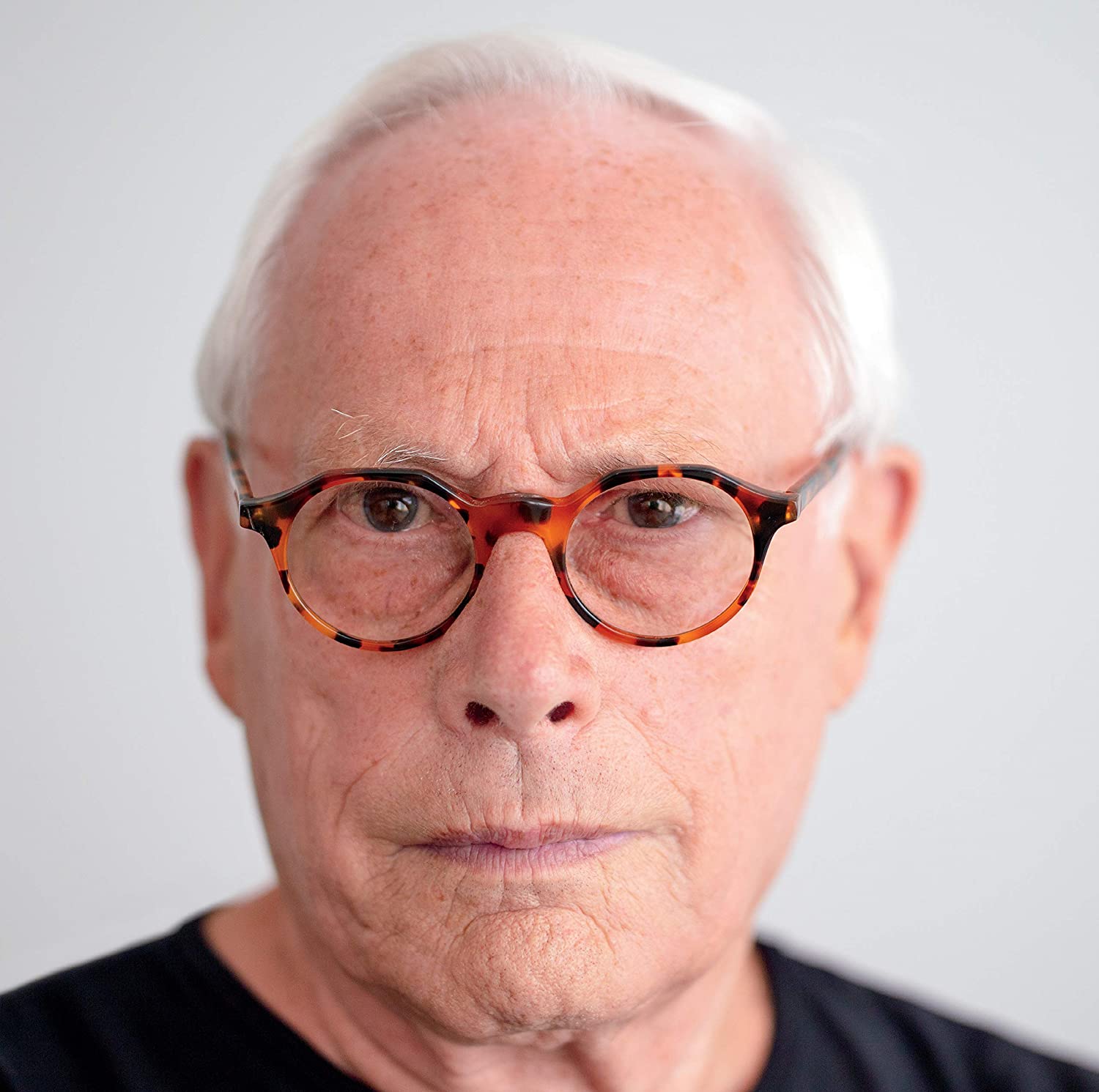 A man with white hair and tortoiseshell glasses against a plain white background.