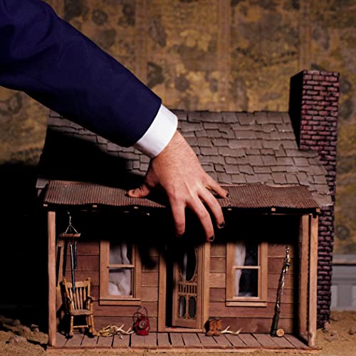 A person in a blue suit reaches down to touch the roof of a miniature cabin.