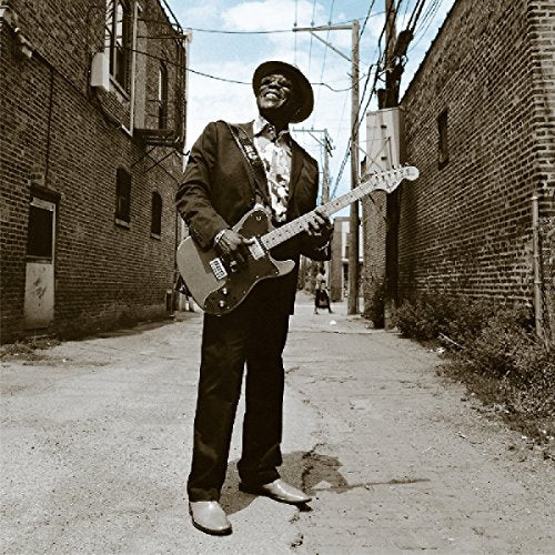 Buddy Guy smiling while playing an electric guitar in a brick alleyway.