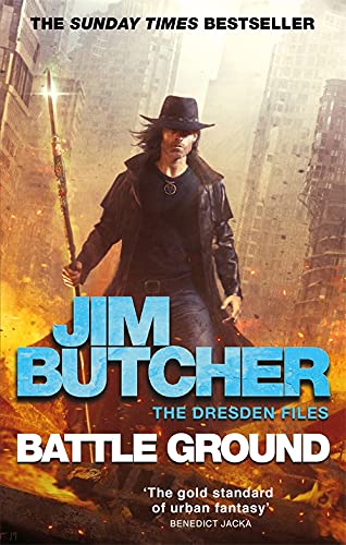 A man in a hat and duster holds a staff amidst a fiery urban battlefield.