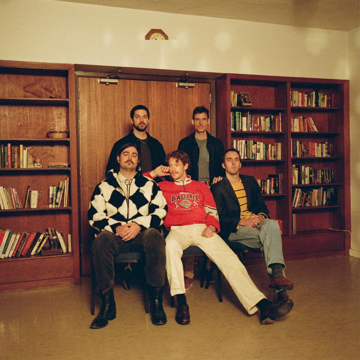 Caamp band portrait, five men seated in front of bookshelves.