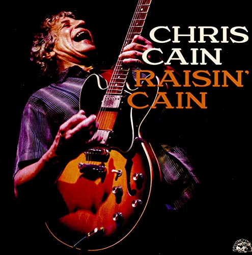 Chris Cain laughing while playing a hollow-body electric guitar against a black background.