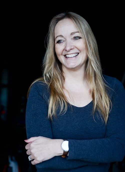 A smiling blonde woman with arms crossed, wearing a dark blue long-sleeved shirt.
