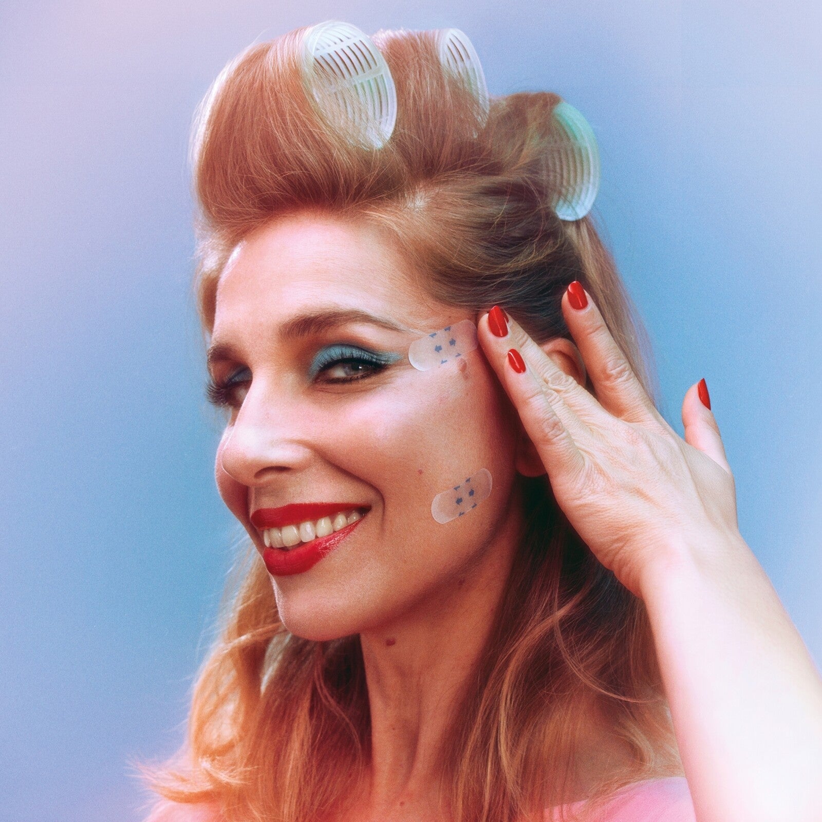 Woman with red hair in rollers, applying stickers to her face.