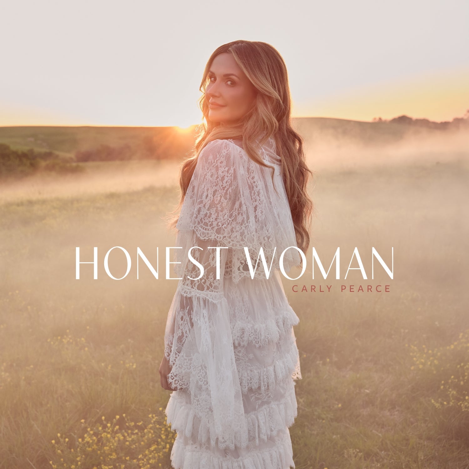 Carly Pearce in a white lace dress standing in a misty field at sunset.