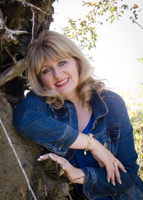 Woman with blonde hair smiling, leaning against a tree branch.