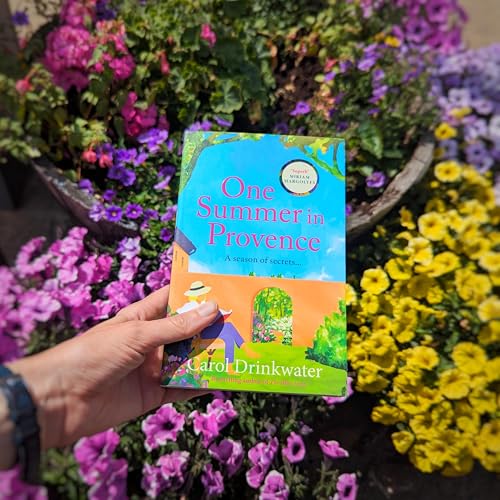 A hand holds the book One Summer in Provence in front of colorful flowers.
