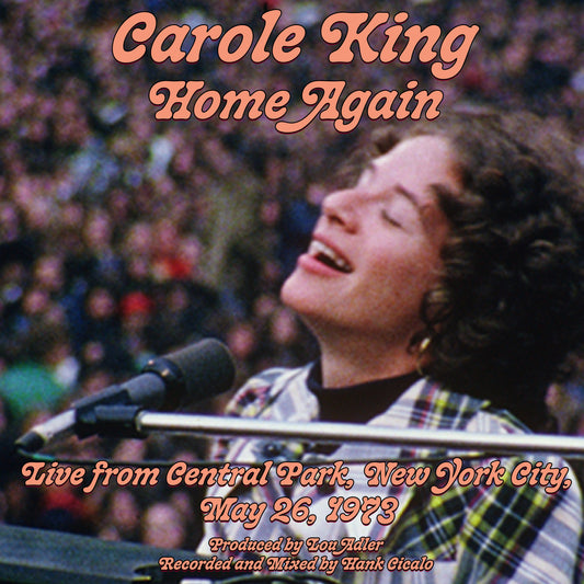 "Carole King sings into a microphone, with a crowd in the background."