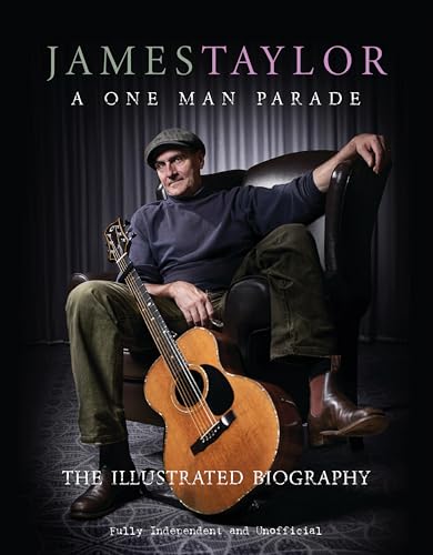 James Taylor wearing a cap and holding an acoustic guitar while sitting in chair.