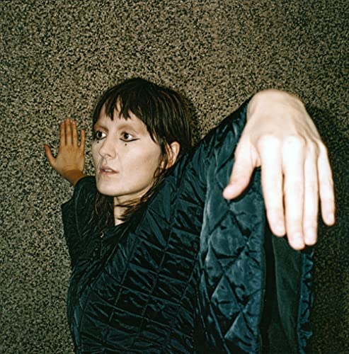 Cate Le Bon poses in a dark quilted jacket against a textured wall.