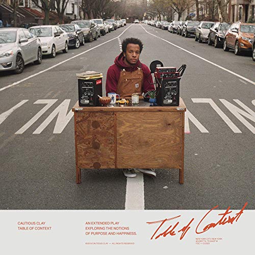 "Man in a red hoodie stands behind a wooden cart on a city street."