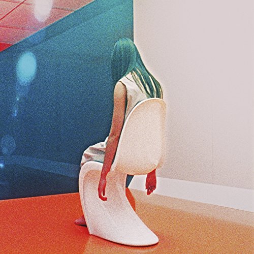 A person with long hair sits on a white chair against a blue wall.