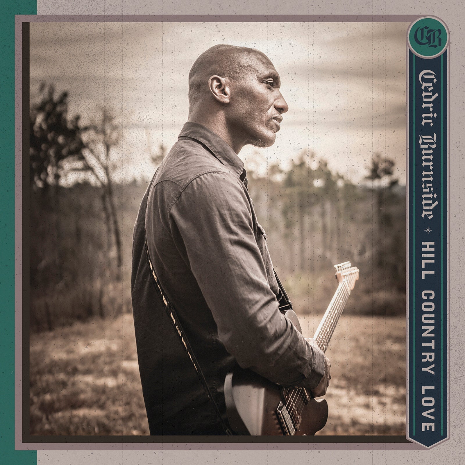 Cedric Burnside in profile holding an electric guitar in a field with trees.