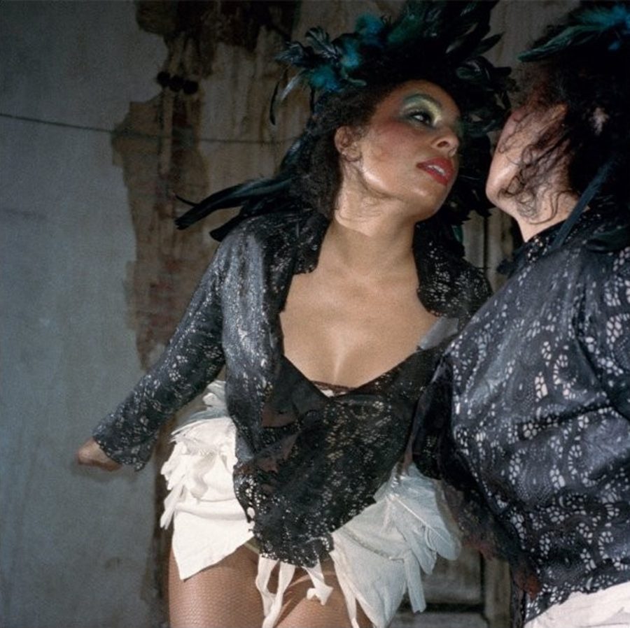 Celeste, wearing lace and feathers, poses against a textured wall.