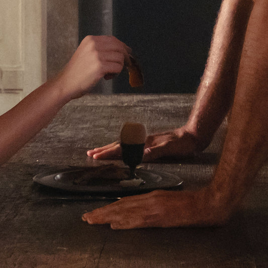 A hand holds a piece of food above an egg cup on a wooden table.