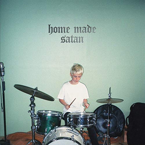 Blond child playing drums in a room with a green wall and drum set.