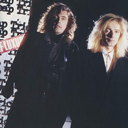 Two men with long hair wearing dark suits pose against a patterned background.