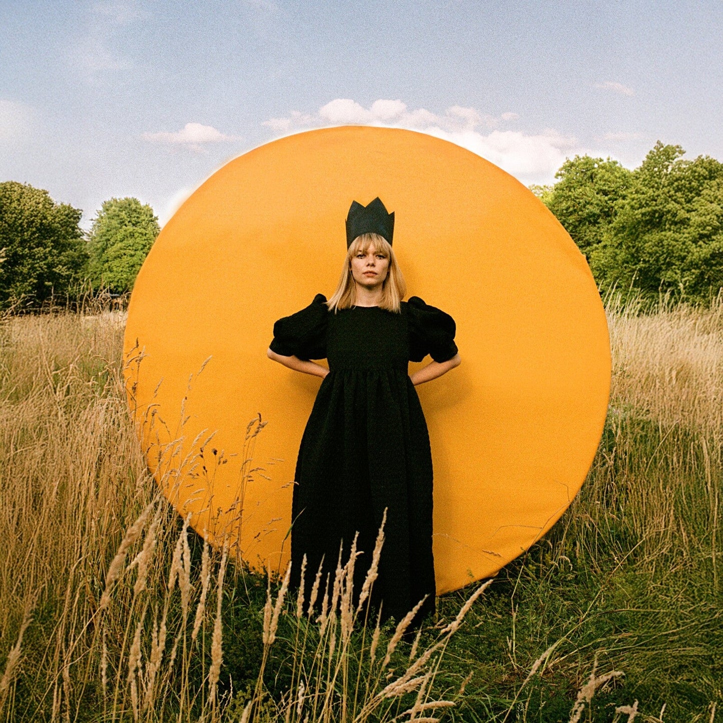 Woman in black dress and crown stands before a large yellow circle.