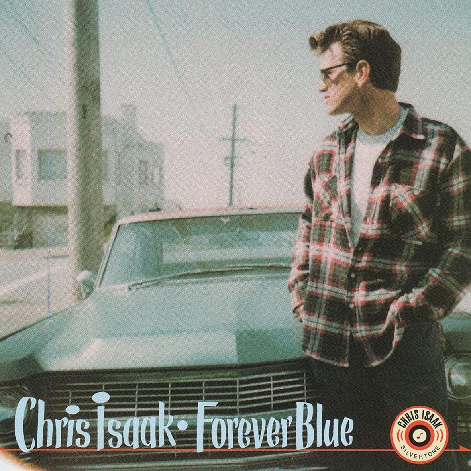 Chris Isaak in glasses and plaid, leaning on a silver car.