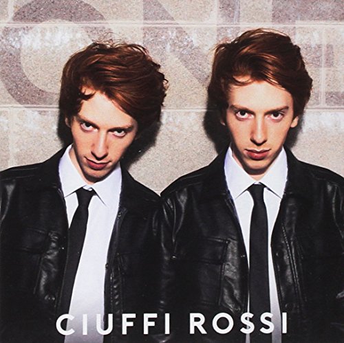 Two men in black jackets and ties stand against a brick wall background.