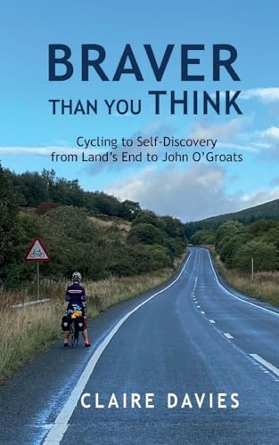 A cyclist stands on the shoulder of a road with hills and sky above.