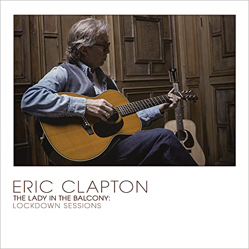 Eric Clapton seated, playing an acoustic guitar against a wooden door.