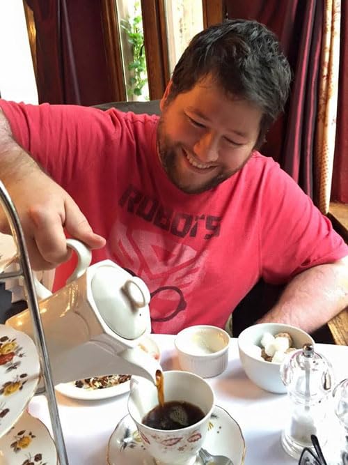 A smiling man in a red t-shirt pours tea into a teacup at a table.