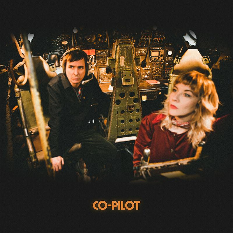Two people sit in a cockpit with vintage controls and lighting.
