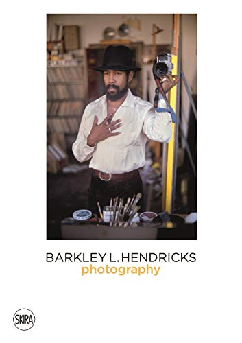Barkley Hendricks in hat and white shirt, holding a camera.