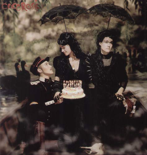 Three people in dark Victorian clothing hold black umbrellas over a lit birthday cake.