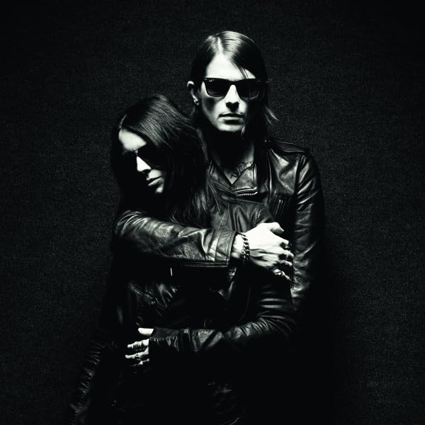 Two people in leather jackets and sunglasses embrace against a dark background.